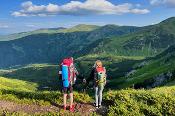 Naklejka premium Back view of two hikers, holding hands, stand on trail overlooking mountain landscape. Tourists equipped with backpacks and trekking poles. Rolling hills, rocky outcrops, bright blue sky with clouds.