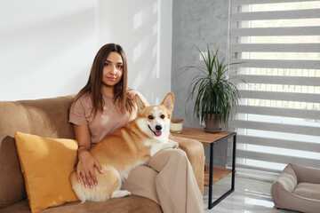 Joy emanates from a woman playing with her corgi dog on a sofa. Sunlight filters in as she tickles...