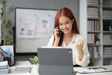 Obraz premium Excited asian businesswoman working in the office using a tablet and talking on the phone while raising her fist celebrating success