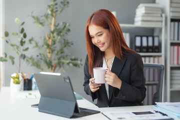 Asian businesswoman smiles while enjoying coffee at work in a modern office, focused on her laptop amidst technology and paperwork, exuding confidence and professionalism