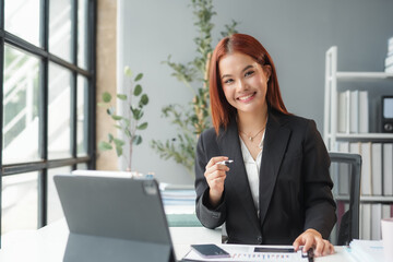 Young asian businesswoman smiling while sitting at her desk in a modern office, working on a laptop computer with paperwork and financial charts