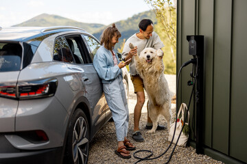 Portrait of a young couple with their cute white dog near house and modern electric car outdoors....