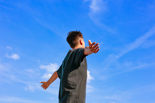 Young teenager boy with arms outstretched breathing in fresh air against blue sky. Teen enjoying open air in nature, healthy good life balance lifestyle. 