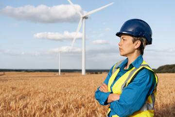Wind energy technician surveys golden fields under clear sky with wind turbines spinning in background during sunny afternoon