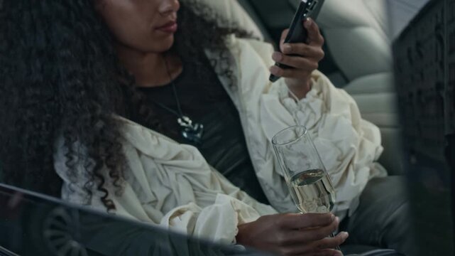 Cool Biracial influencer girl wearing stylish fancy outfit sitting with glass of champagne in back seat of luxury car and scrolling on smartphone