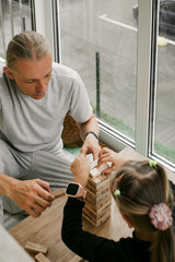 Child and an adult focus on building wooden tower game together at table indoors. Father and daughter enjoying free time. Quality family communication and informal education