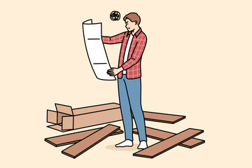 Man assembles furniture with own hands and holds large paper instruction manual, standing among boards. Young guy who assembles furniture is feeling unsure due to lack of experience.