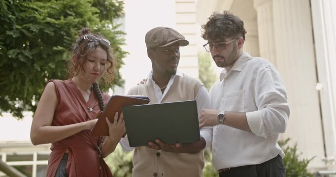 In an outdoor business setting, a diverse group of professionals uses a laptop and tablet to collaborate, highlighting their teamwork and effective use of technology for project success.