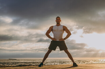 Young strong sporty athletic toned fit sportsman man wear sports clothes stand akimbo, arms on waist warm up training at sunrise sun dawn over sea beach outdoor on pier seaside in summer day morning.