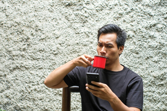 A Southeast Asian man wearing black t shirt sitting on bamboo chair in the terrace, drinking black coffee on red mug and holding smartphone, relax time.