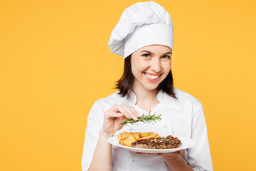 Young chef cook baker woman wearing white shirt black apron uniform toque chefs hat hold in hand plate with dish add rosemary to steak isolated on plain yellow background studio. Cooking food concept.