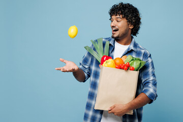 Young happy surprised Indian man in shirt hold brown craft bag for takeaway mock up with food products throw up lemon fruit isolated on plain blue background Delivery service from shop or restaurant