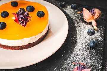 Dessert or cake with layers of chocolate, curd cream, mango jelly and orange, decorated with food dried flowers on a white plate on a black background. Original presentation, food design.