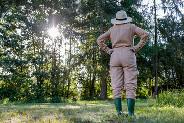 Woman gardener in straw hat and brown garden suit  in the garden on warm autumn sunny day. Lush vegetation and grass and trees background. Autumn gardening.