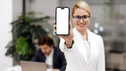 In a modern office, a young woman holds a phone with a white screen in her hands. Ideal for displaying digital advertising, interactive content or promotional messages.