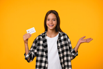 Little kid teenager child school girl wears casual clothes hold in hand mock up of credit bank card isolated on bright orange yellow background. Childhood banking concept