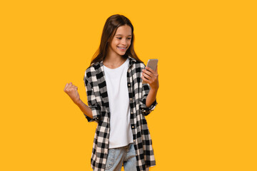 Happy excited like a winner girl teenager student schoolkid holding smart phone for mobile application, laughing, winning a lot of money. Billionaire isolated in yellow background