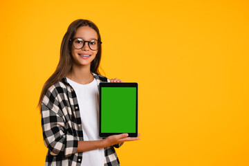 Smart smiling caucasian schoolgirl teenager child pupil student holding digital tablet showing blank screen with advert place mockup isolated in yellow background