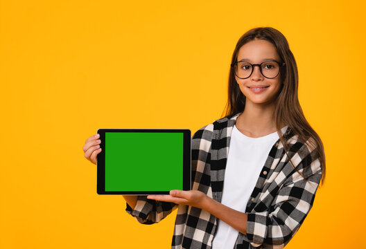 Cutout portrait of smiling caucasian schoolgirl teenager child pupil student holding digital tablet showing blank screen with advert place mockup isolated in yellow background
