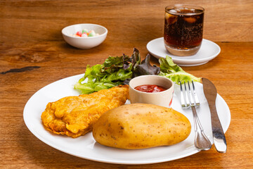 Closeup view battered fried fish, boiled potato, fresh green lettuce, a cup of ketchup, metal fork and table knife in white ceramic plate with glass of ice cola water on wooden table.