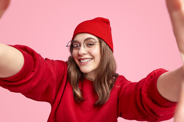 Cheerful young woman in red beanie and sweater taking a selfie against a pink background, expressing joy and confidence.