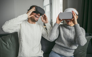 Obraz premium Happy couple using virtual reality headsets and experiencing immersive technology while sitting on a couch at home.