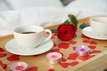 Wooden tray with burning candles, rose, cups of coffee and red paper hearts on bed indoors, closeup