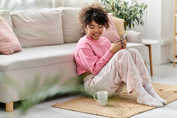 A young woman in pink loungewear smiles while using her phone at home.