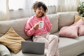 A young woman laughs on the couch while sipping her favorite beverage.