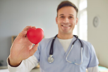 Portrait of smiling friendly male doctor cardiologist holding red heart in his hands in clinic and looking at camera in medical office. Medicine, people, health care and cardiology concept.