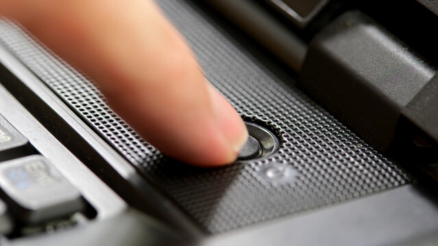 Finger pressing a generic round dark black power button on a simple office laptop, closeup detail, one person. Man turning on a laptop computer, or turning the device off resetting it abstract concept