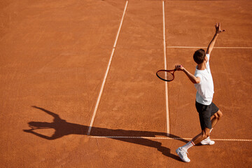 Back view image of focused young man in white t-shirt and black shorts, tennis player training on clay court. serving ball with racket. Concept of sport, competition, active and healthy lifestyle