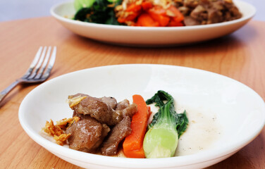 On a divided plate, there was pork stir-fried with sauce and chopped carrots, whole vegetables, and fried garlic on the side. Next was a large plate placed on a wooden table.

