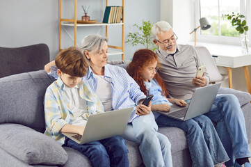 Happy grandparents with grandchildren girl and boy sitting on sofa using modern laptop and...