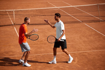Two emotional happy men, tennis players in motion on clay court showing support and respect to each other, shaking hands after golf game. Concept of sport, competition, active and healthy lifestyle