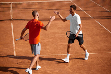 Two smiling men, tennis players shaking hands on clay court after competitive match, showing mutual respect and support. Concept of sport, competition, active and healthy lifestyle