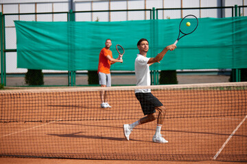 Two men, friends spending active time on outdoor tennis court, playing tennis, growing game skills and endurance. Concept of sport, competition, active and healthy lifestyle