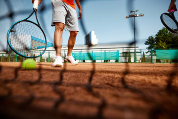 Image taken near the net. Focus on male legs, tennis player with racket on clay outdoor court during intense summer game. Concept of sport, competition, active and healthy lifestyle