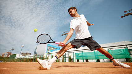 Dynamic image of focused young man in white t-shirt playing tennis on outdoor clay court, showing energy and focus during match. Concept of sport, competition, active and healthy lifestyle