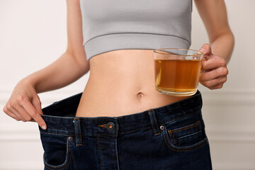 Weight loss concept. Woman with cup of diet tea and big pants near white wall, closeup