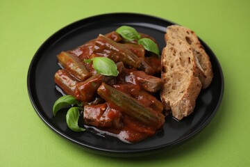 Tasty stew with okra, tomato sauce, bread and basil on light green background, closeup