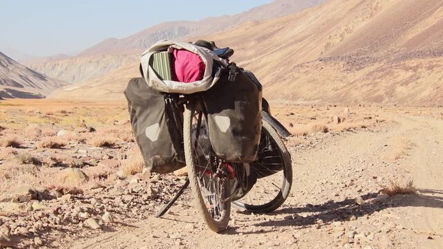 Red touring bicycle loaded with heavy gear in four pannier bags stand on side of gravel road surrounded by wild arid Pamir mountains alpine nature outdoors. Solo adventure travel