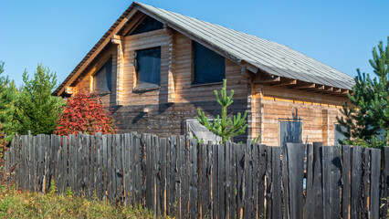 Obraz premium A wooden timber house under construction is located behind a wooden fence. Construction of a country house made of timber. There are trees growing near the house made of timber.