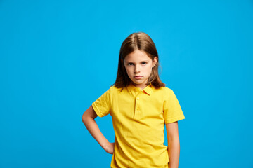 Portrait of teen girl in yellow polo shirt standing with serious face, expressing dissatisfaction against blue studio background. Concept of childhood, kids emotions, lifestyle