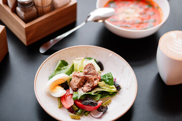 Meal setup on a table with salad, meat slices, half-cut boiled egg, soup with herbs, and coffee with latte art dark surface with utensils and wooden box in the background top-down angle medium plan
