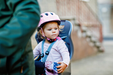 A smiling toddler girl wearing a pink helmet sits securely in a bicycle child seat, ready for a ride. The background is blurred, focusing on the child joyful expression