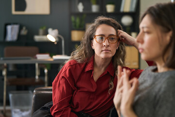 A counselor, wearing glasses and red shirt, is attentively listening to client in well-organized...