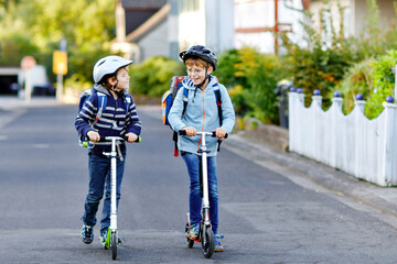Two school kid boys in safety helmet riding with scooter in the city with backpack on sunny day....