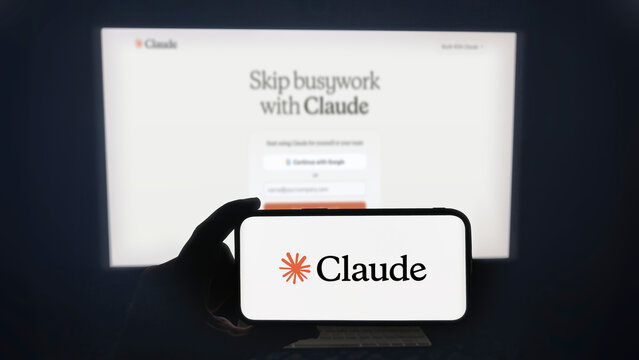 Claude app running on a modern mobile phone. The smartphone is being held in landscape mode, in the darkness. A PC monitor with the Claude desktop application in the background