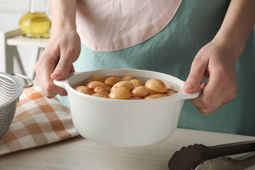 Woman holding pot with raw potatoes at white wooden table, closeup